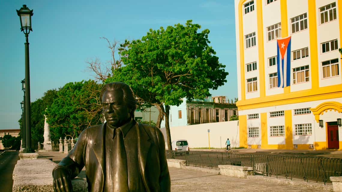 Estatua de Nicolás Guillén en la Alameda de Paula, al fondo un edificio colonial exhibe la bendera de Cuba