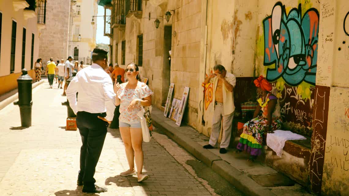 Habaneros conversan en la Calle Empedrado, al fondo cuadros en venta para turistas