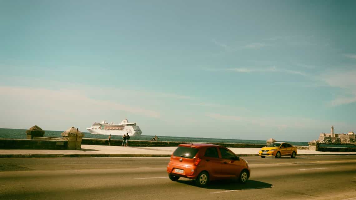 Coche y taxi recorren el Malecon, al fondo barco crucero se aleja del Faro del Morro en la entrada de la Bahia de La Habana