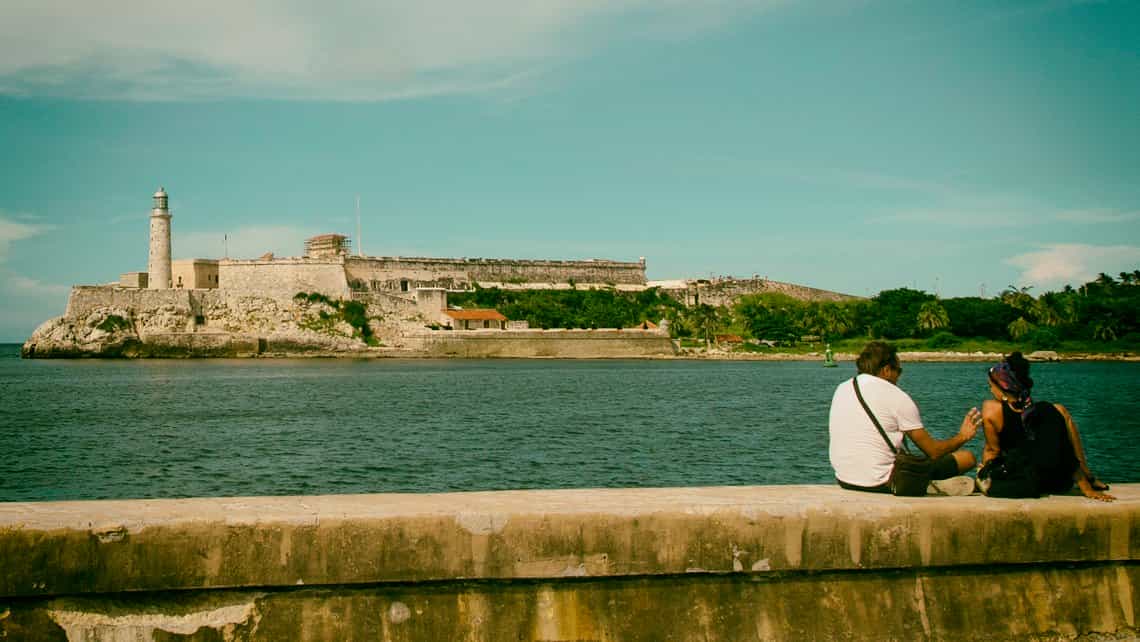 Pareja de enamorados conversa sentados en el muro del Malecon, al fondo el Faro del Castillo del Morro