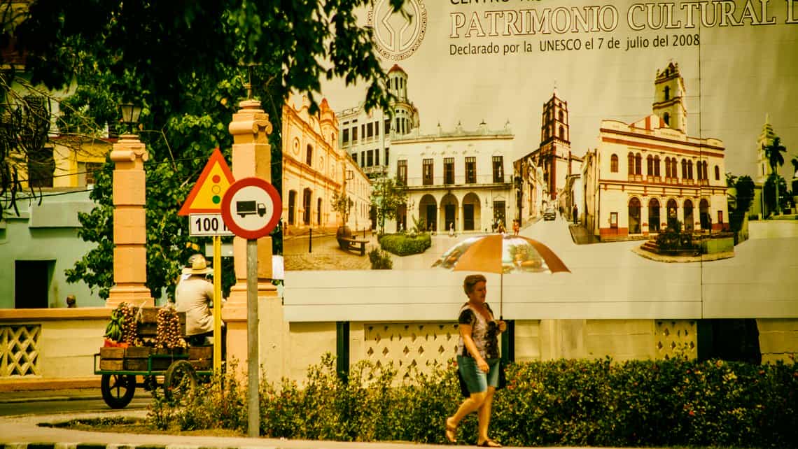 Senora recorre las calles de Camaguey en dia soleado, al fondo cartel celebra la declaracion de la ciudad como Patrimonio Cultural de la Humanidad por la UNESCO