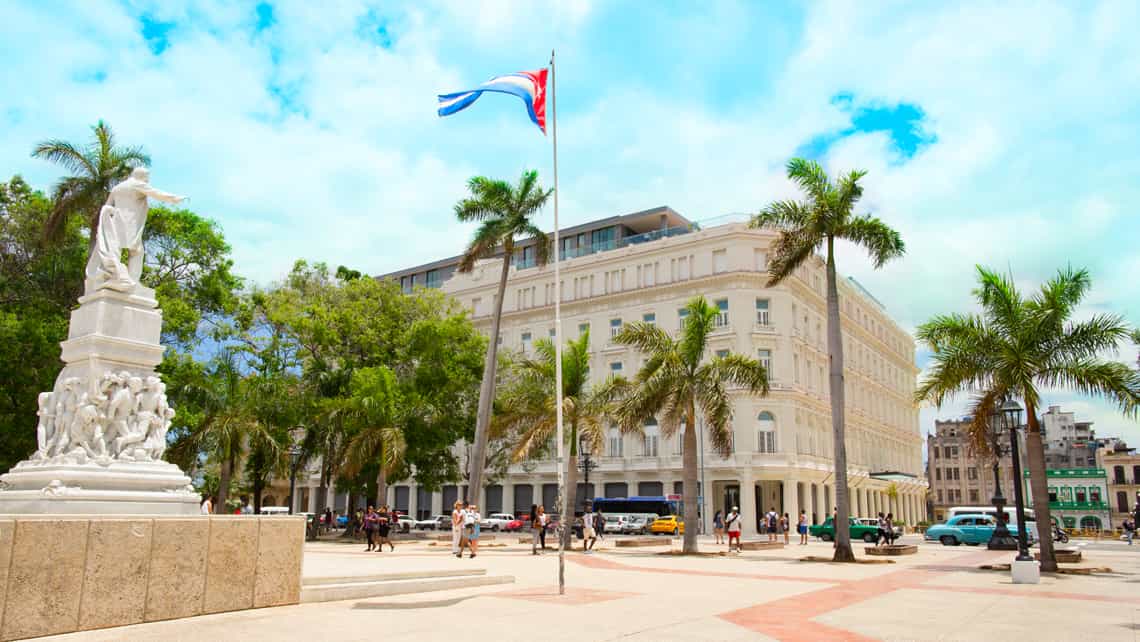 Estatua de Jose Marti en el Parque Central de La Habana, al fondo ondea la bandera cubana y se ve la fachada del Hotel Kempinski Manzana de Gomez